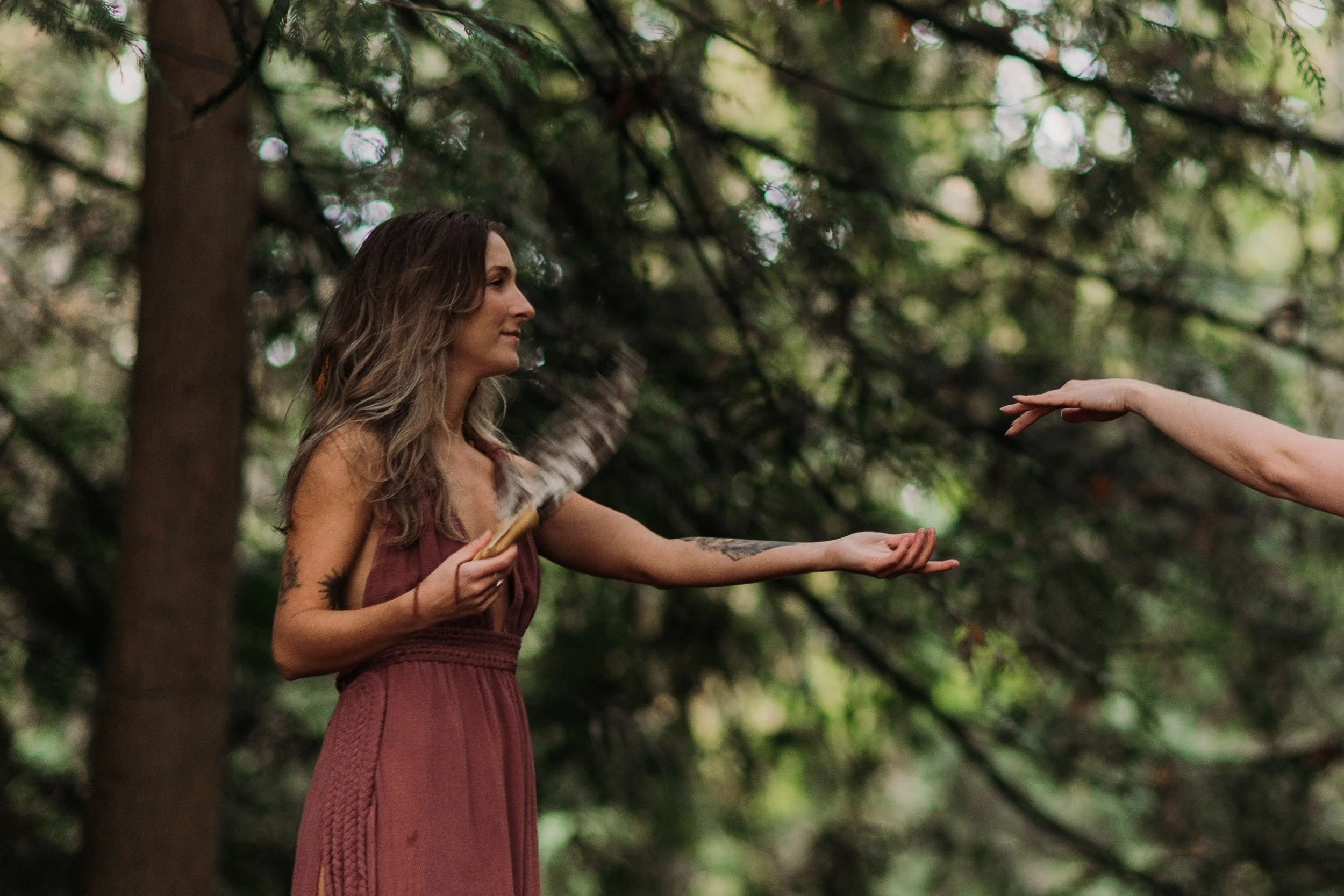 A woman with long, wavy hair and tattoos on her arms is outdoors, holding a smudge fan and reaching out with her hand towards another person whose hand is extended toward her. The background consists of blurred green trees and foliage.