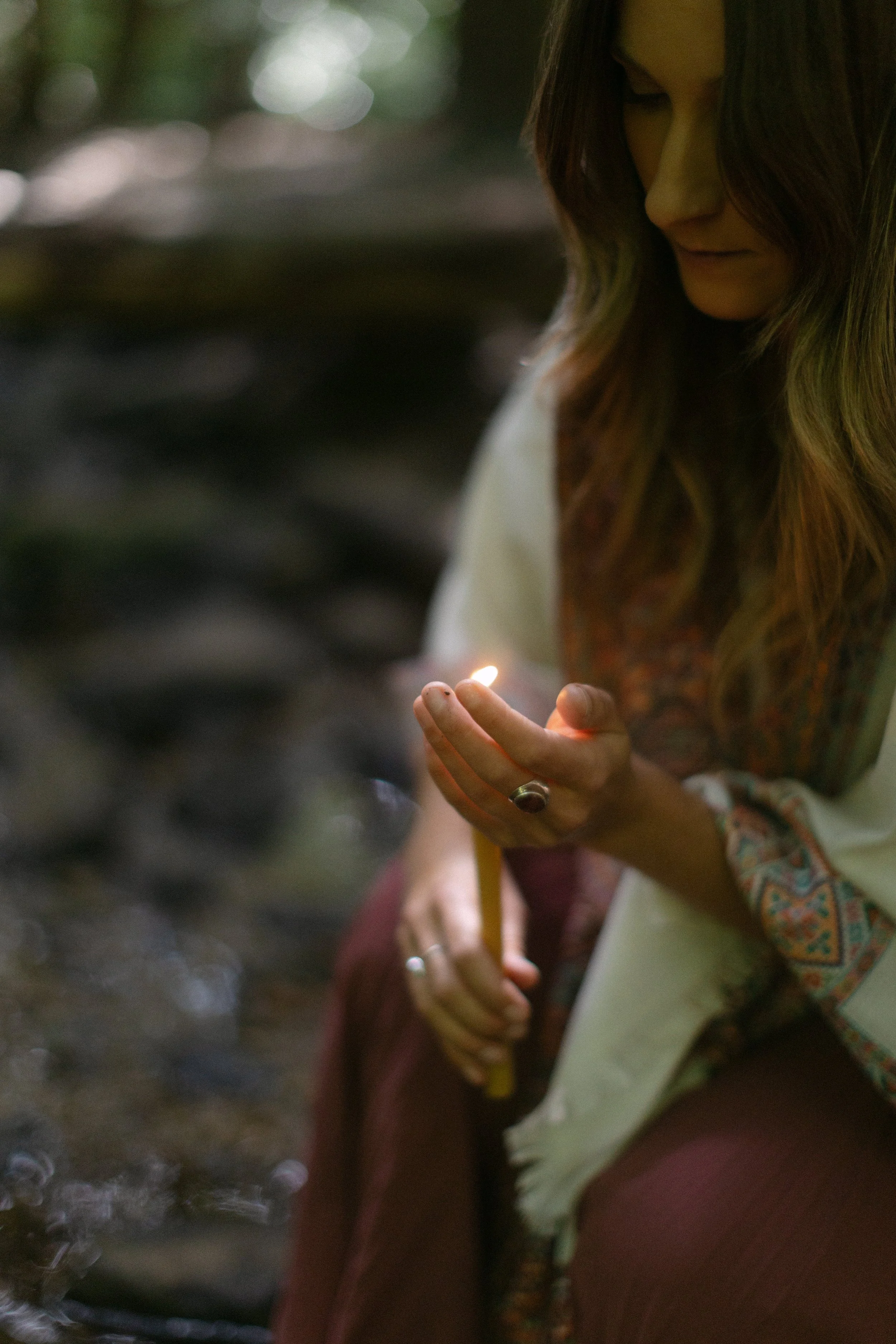 A woman with long hair, wearing traditional clothing, is holding a small lit candle or incense stick, with her eyes closed or looking down, in a serene setting.