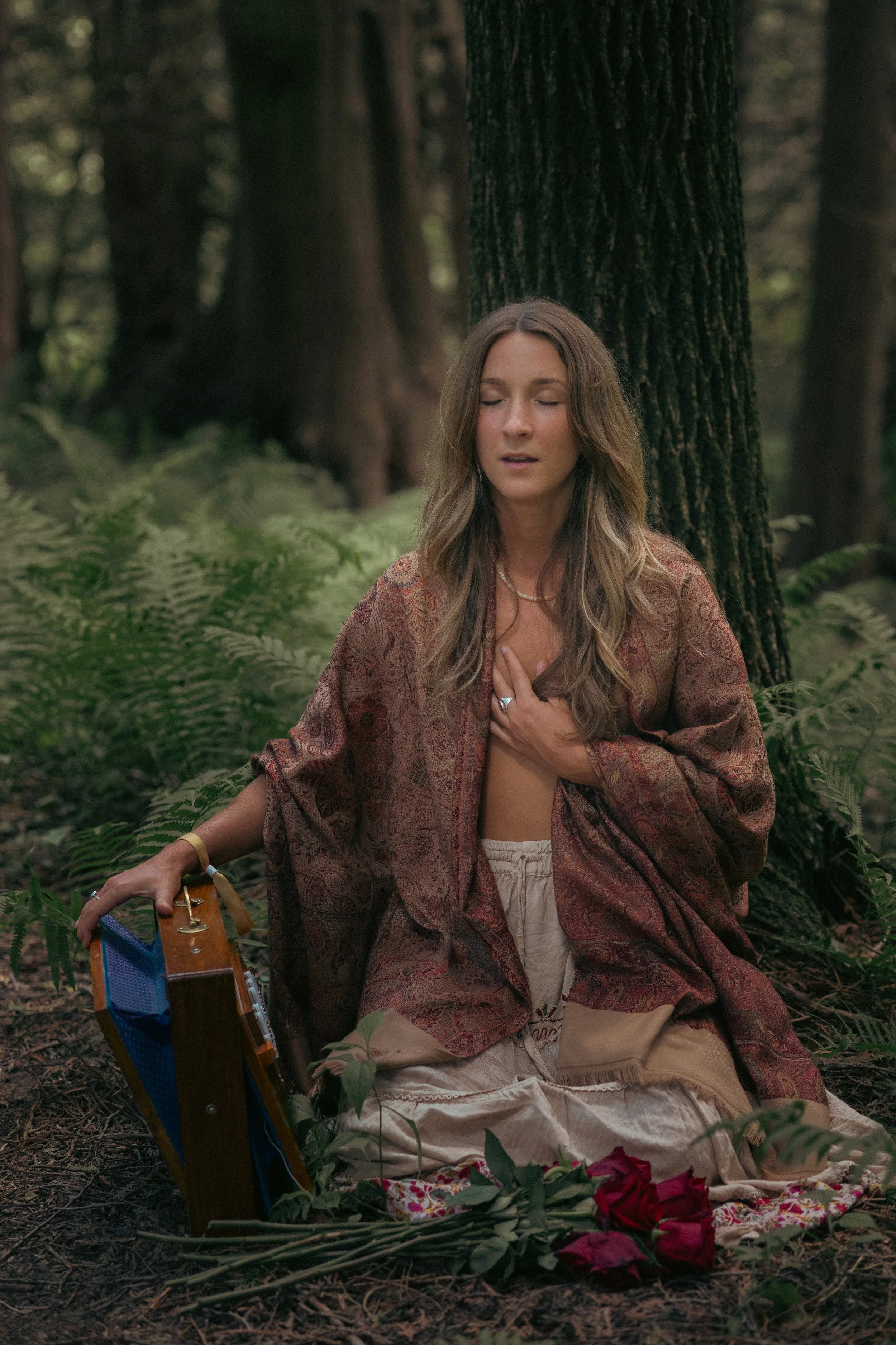 A young woman with long, wavy hair, closed eyes, and a serene expression, sits cross-legged in a forest. She holds her hand to her chest, wearing a patterned shawl, and is surrounded by greenery and fallen red roses. There is a small wooden box with a woven handle next to her.