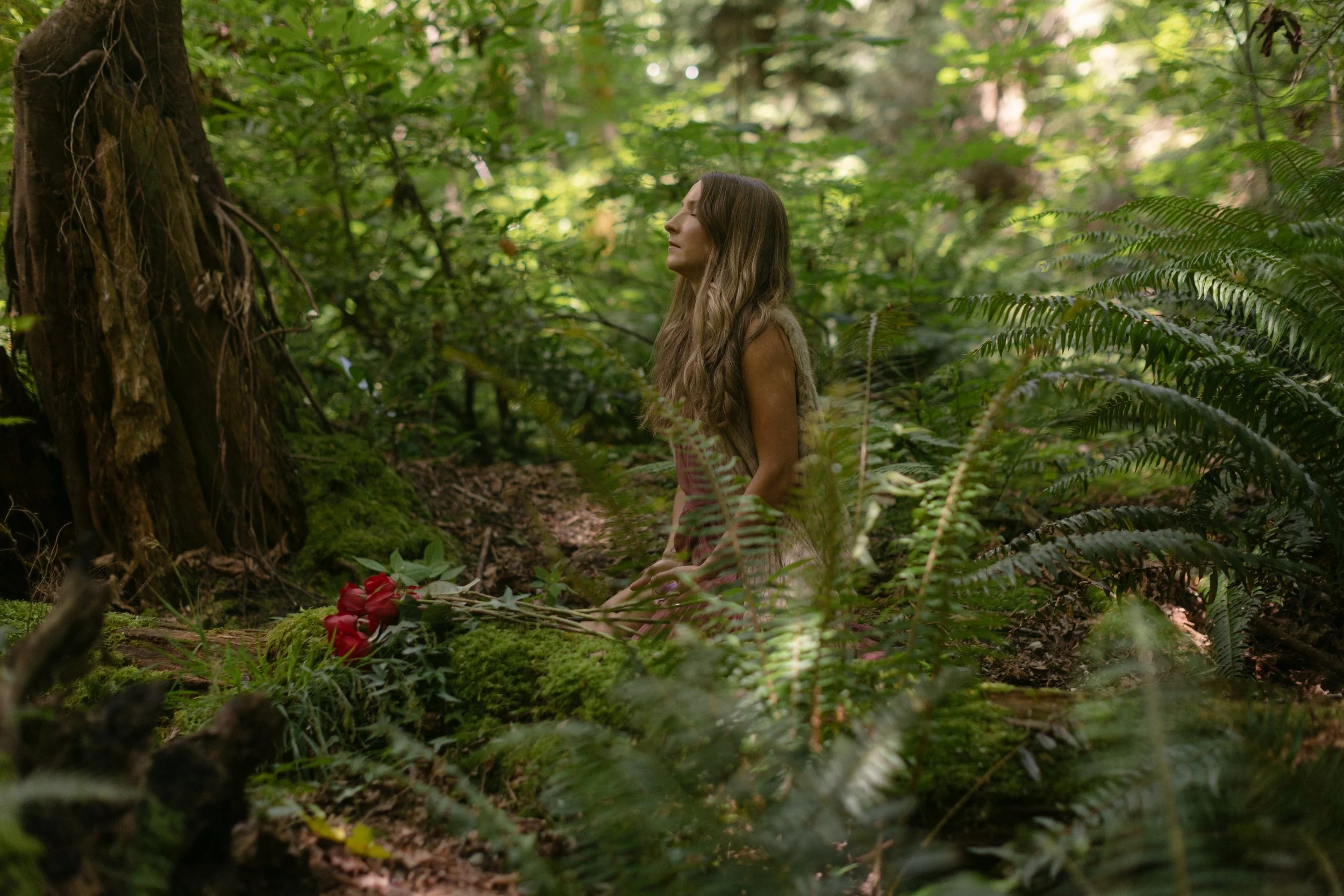 A woman with long brown hair sitting cross-legged in a lush green forest, with trees and ferns surrounding her and a bouquet of red flowers on the ground in front of her.