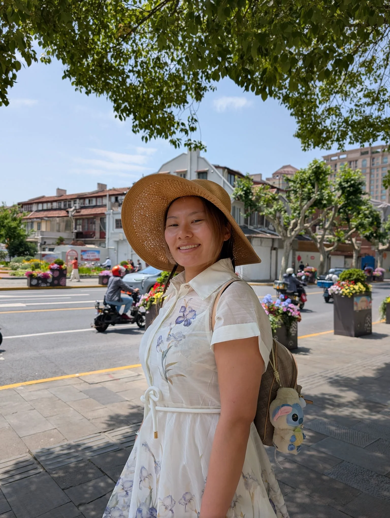 Una donna che indossa un cappello di paglia e una giacca chiara, sorridente, in una via urbana circondata da alberi e piante con fiori, con edifici sullo sfondo e veicoli in movimento.