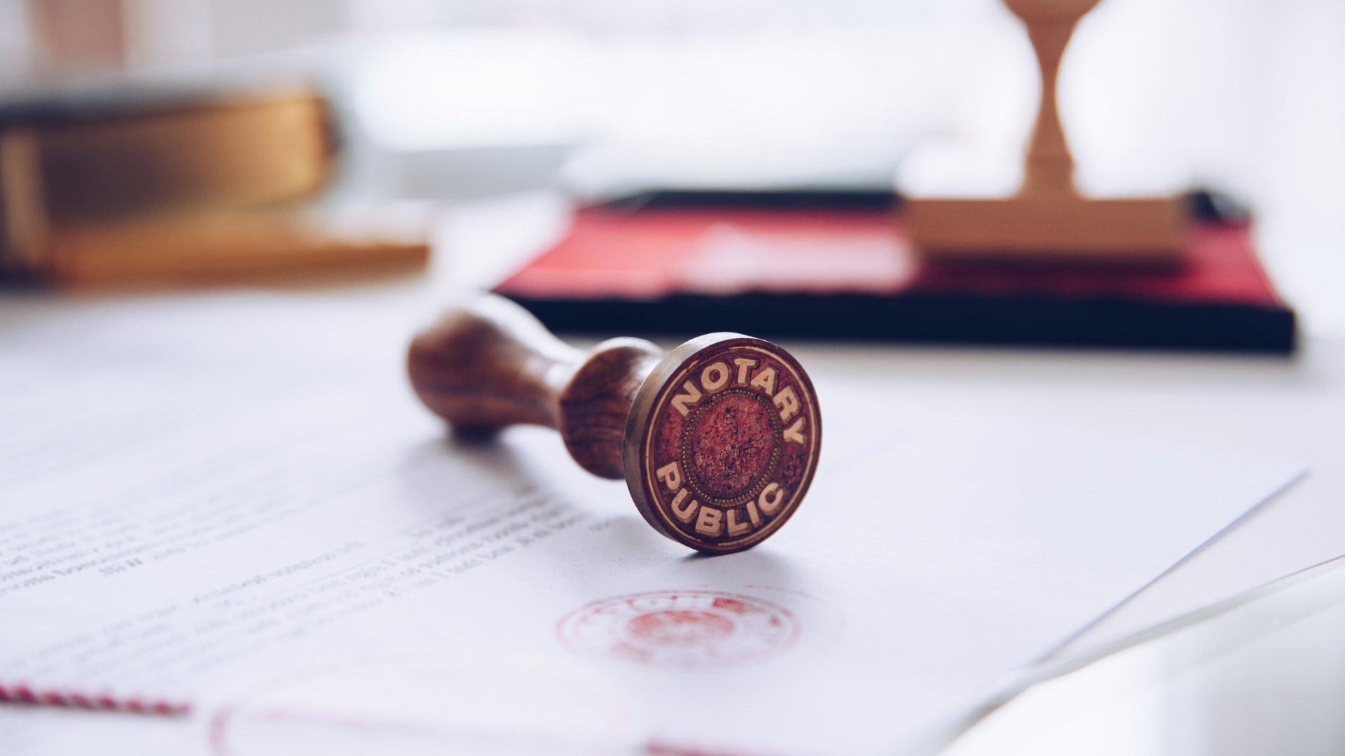 Close-up of a legal rubber stamp with the words "Notary Public" on its side, resting on documents on a desk with books and a blurred background.