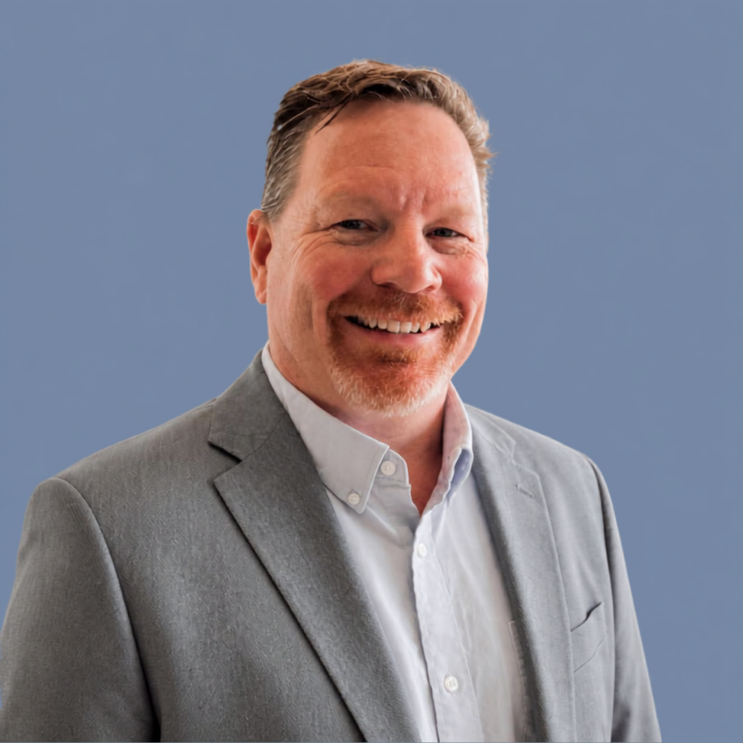 A smiling man with reddish hair and a goatee, wearing a gray blazer and white shirt, standing against a blue background.