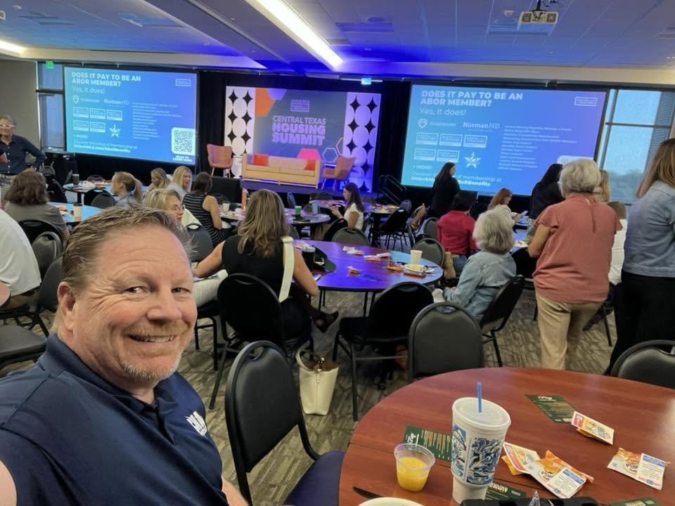 A man taking a selfie at the Central Texas Housing Summit, with a conference room filled with attendees seated at tables in the background. The stage has a large screen displaying event information.
