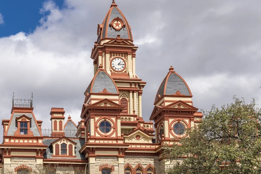 A historic Victorian-style building with a clock tower, pointed roofs, and ornate architectural details, set against a partly cloudy sky with trees in the foreground.