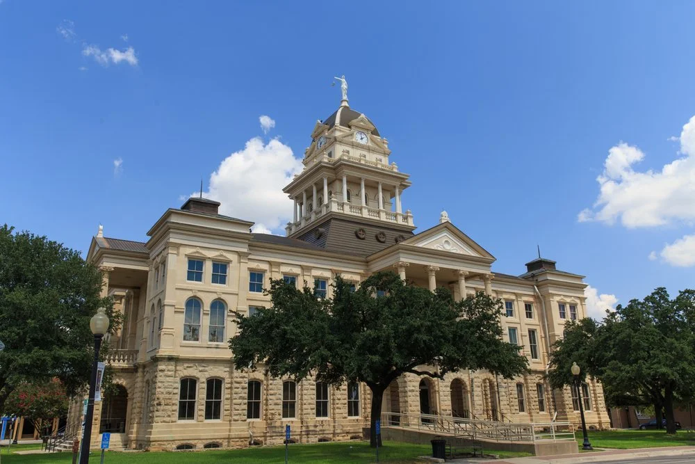 Historic courthouse with clock tower, surrounded by trees, under a blue sky with clouds.