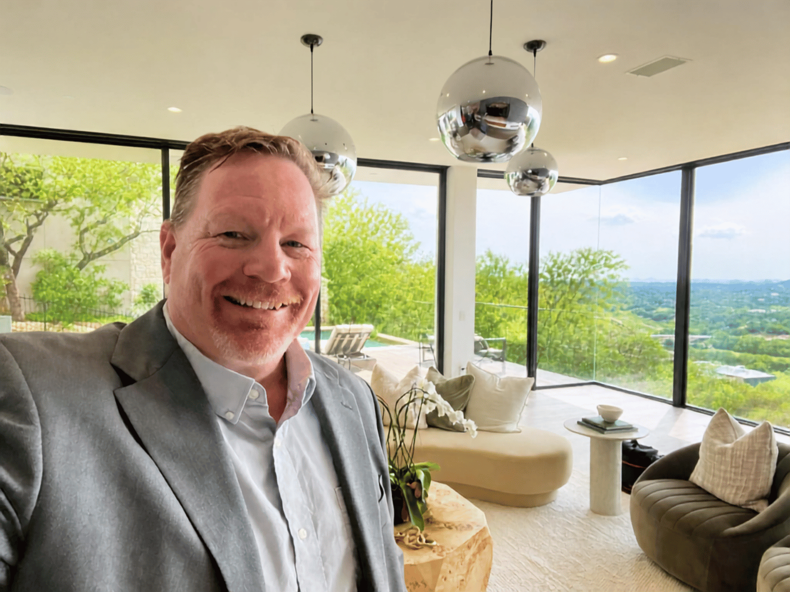 A smiling man with light brown hair and a gray blazer standing in a modern living room with large windows showing a scenic outdoor view.