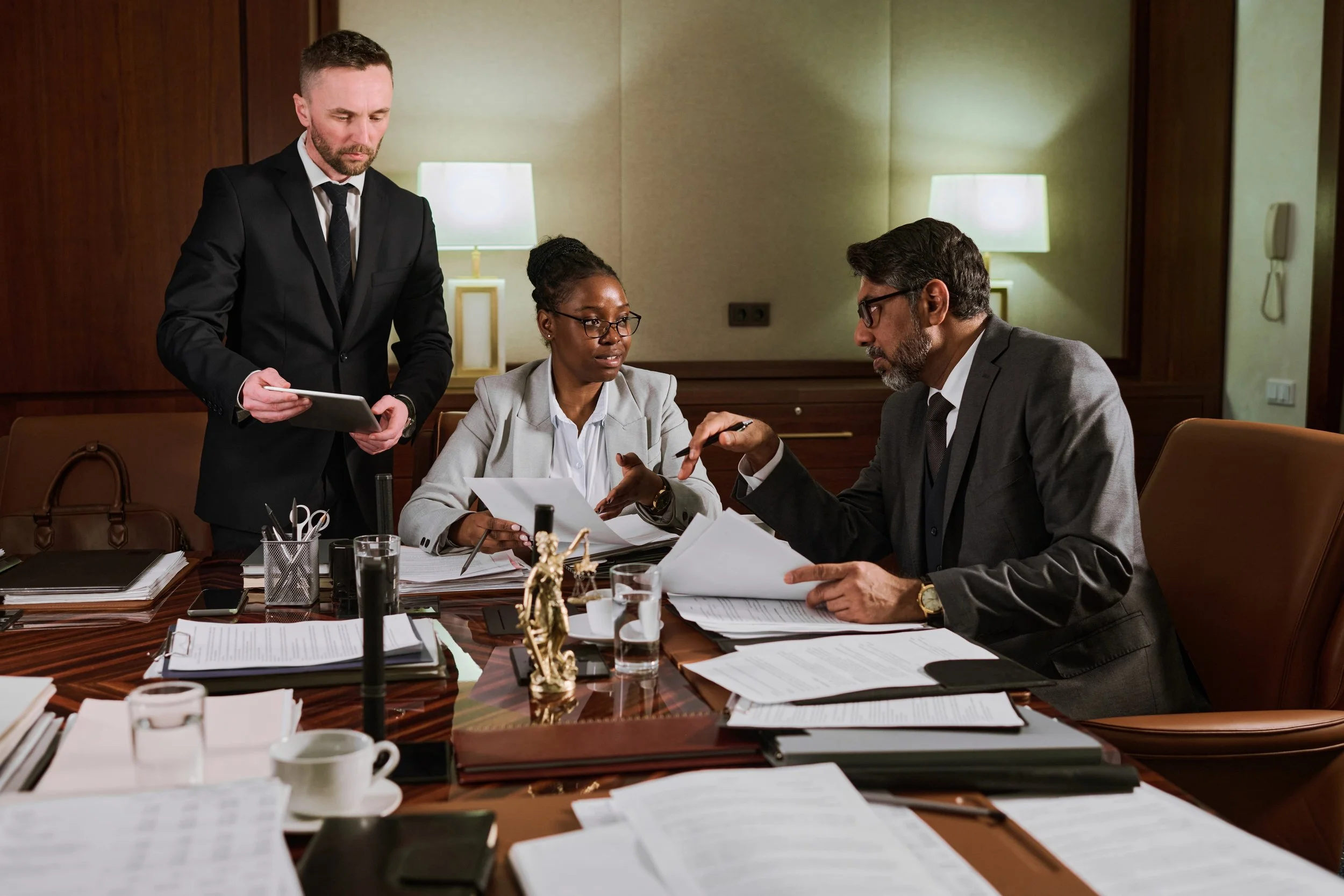Three business professionals engaged in a serious discussion in a conference room. Two men and one woman are surrounded by papers, documents, and office supplies on the table.