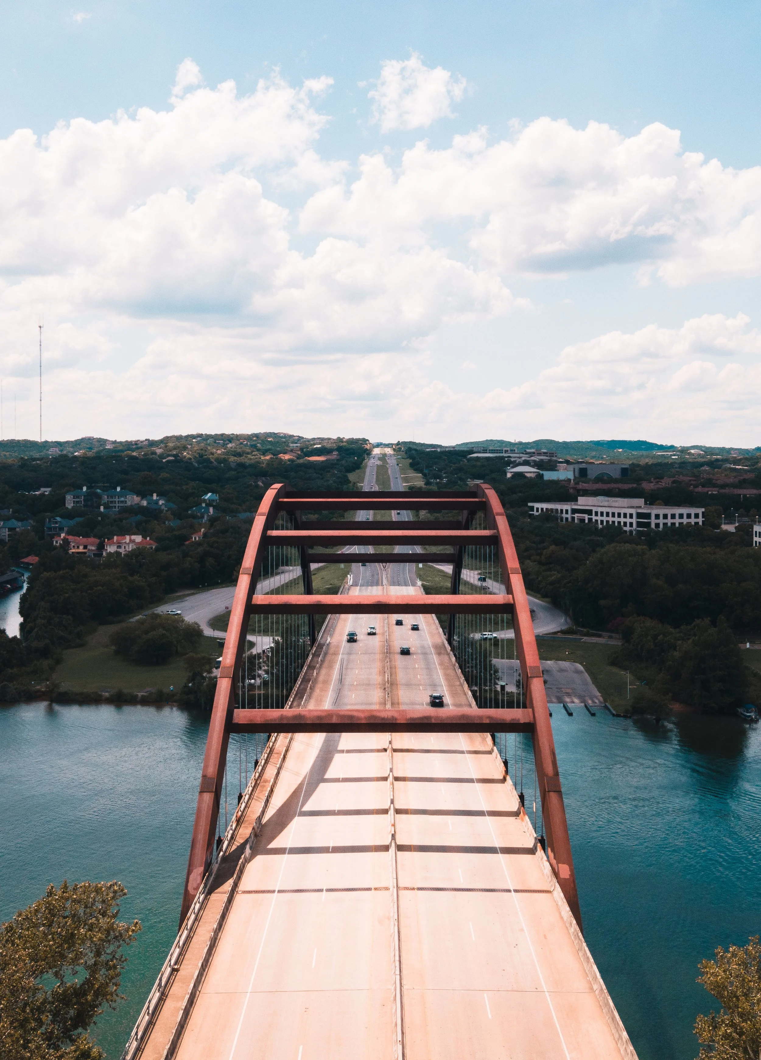 Aerial view of a bridge over a body of water with cars driving in both directions. The bridge has a distinctive arch structure painted in a rust color. In the background, there is a landscape with roads, trees, buildings, and hills, under a partly cloudy sky.