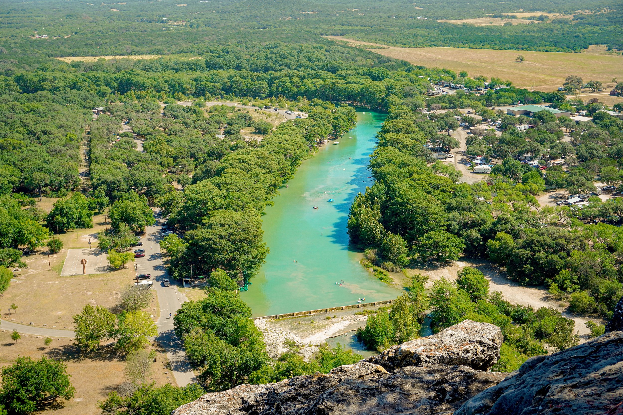 An aerial view of a river with greenish water flowing through a lush, green forest. There are parking lots and roads surrounding the forest, with recreational boats on the river. Farmland and open fields are visible in the background.