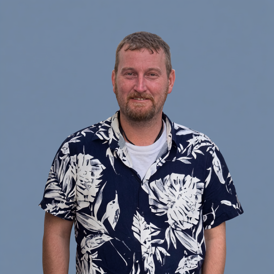 A man with short, light brown hair, a beard, and a smile, wearing a white T-shirt under a navy blue Hawaiian shirt with white leaf patterns, standing against a plain light blue background.