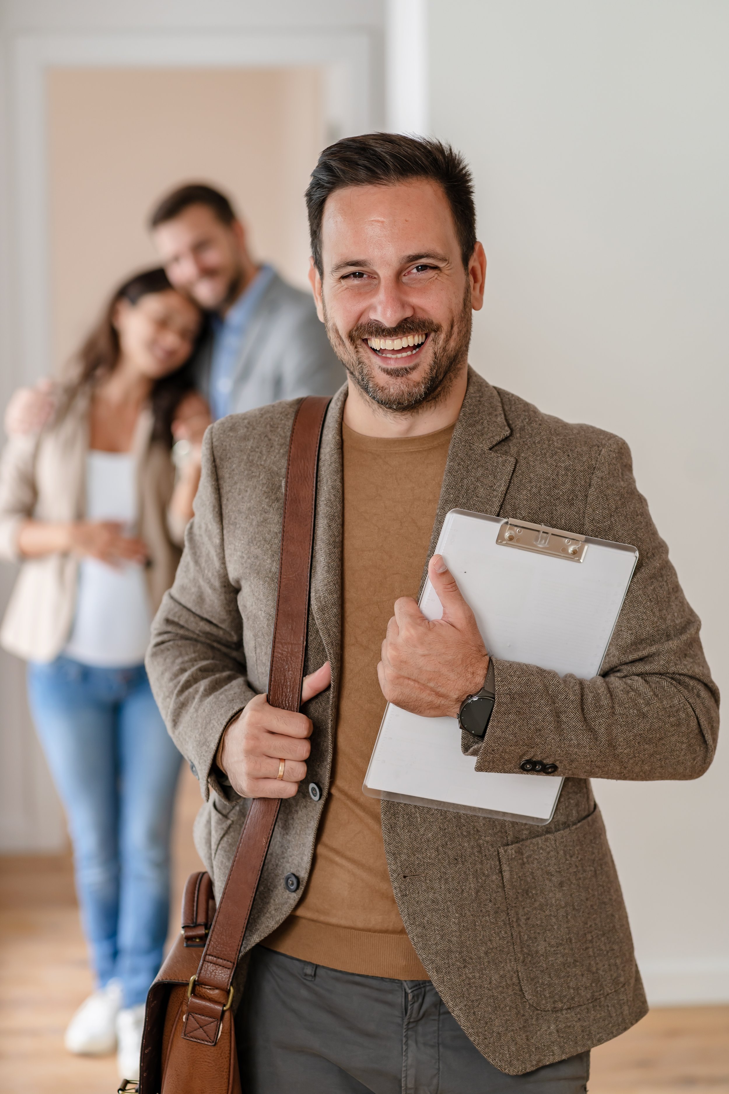 A man in a brown blazer holding a clipboard and smiling. Two people in the background are slightly out of focus, also smiling, in an indoor setting.