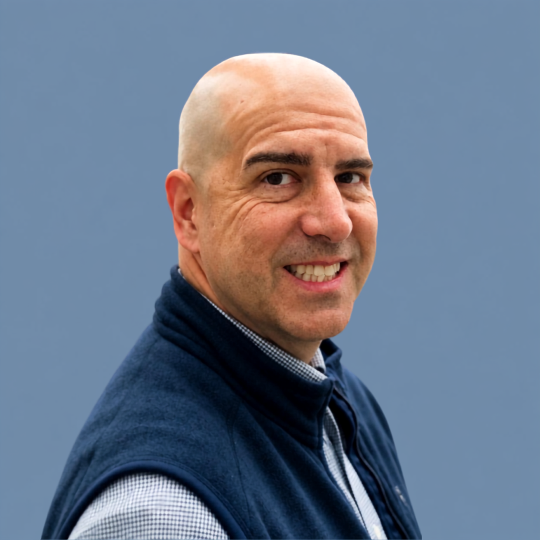 Portrait of a bald man with a light complexion, smiling, wearing a dark blue vest over a collared shirt, against a solid blue background.