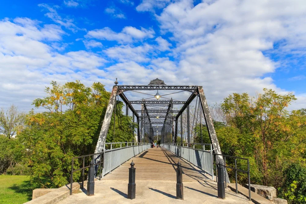 A historic iron bridge with a wooden walkway, surrounded by green trees and a blue sky with scattered clouds.