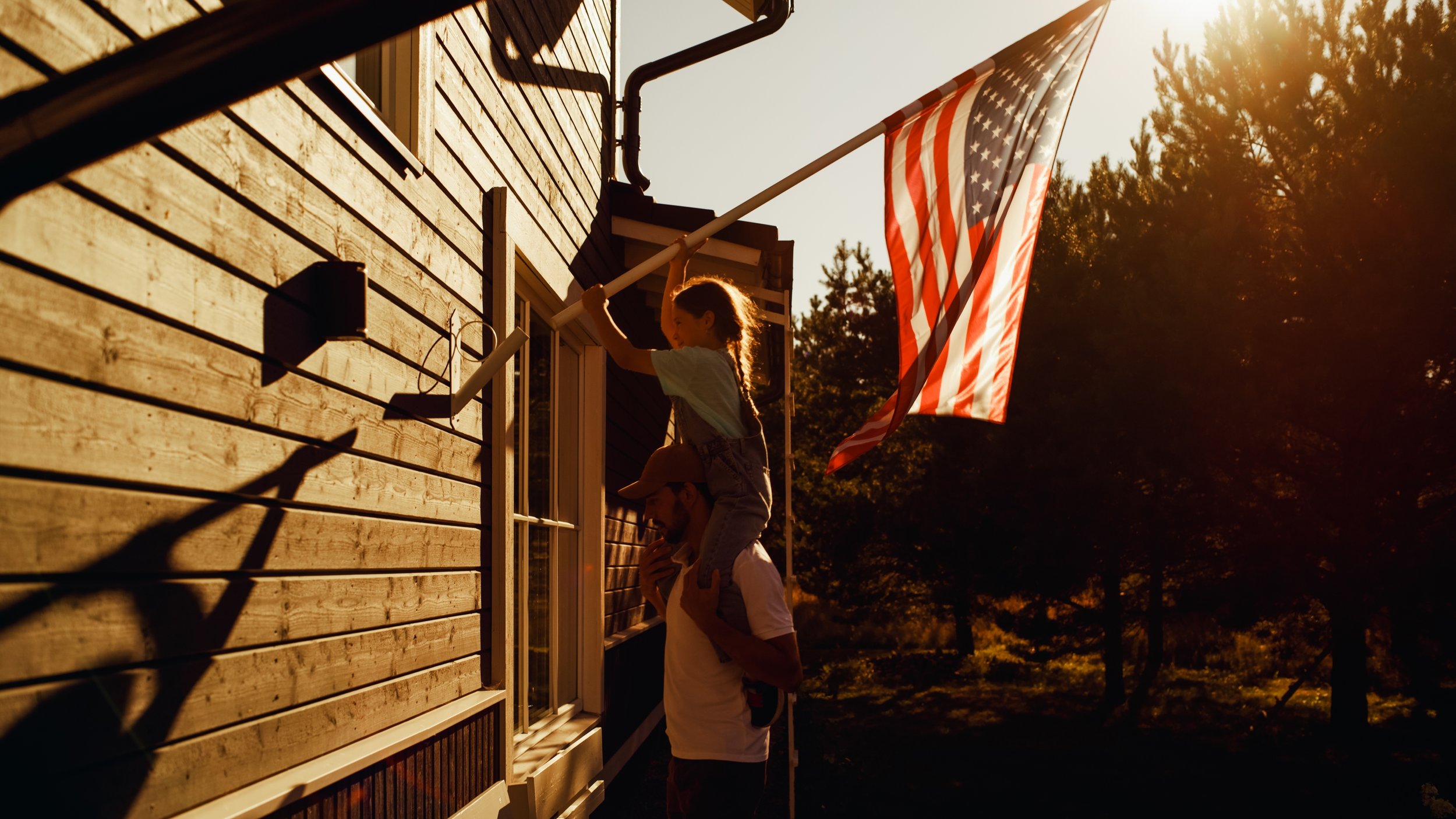 A child and an adult raising an American flag on a flagpole outside a wooden house during sunset.