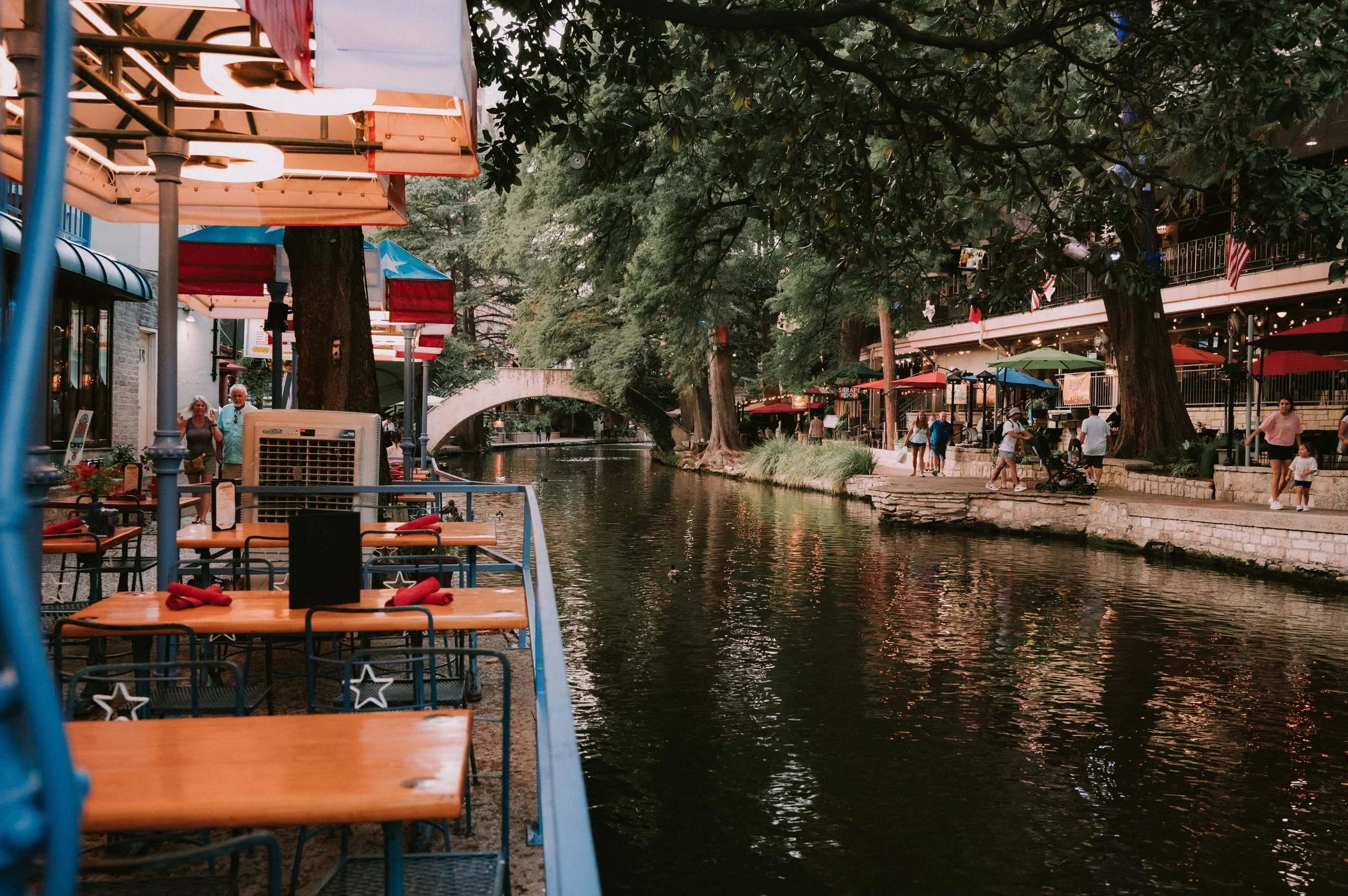Outdoor cafe seating with tables and chairs in front of a canal with trees and people strolling along the sidewalk, some with umbrellas and a bridge in the background.