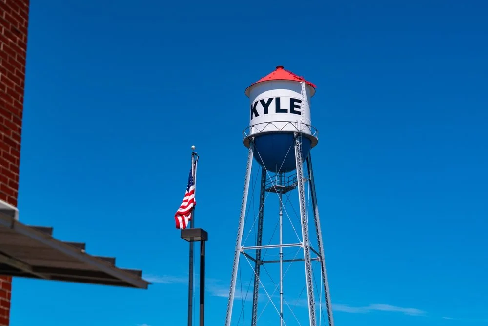 Water tower with the name Kyle written on it, next to an American flag, under a clear blue sky.