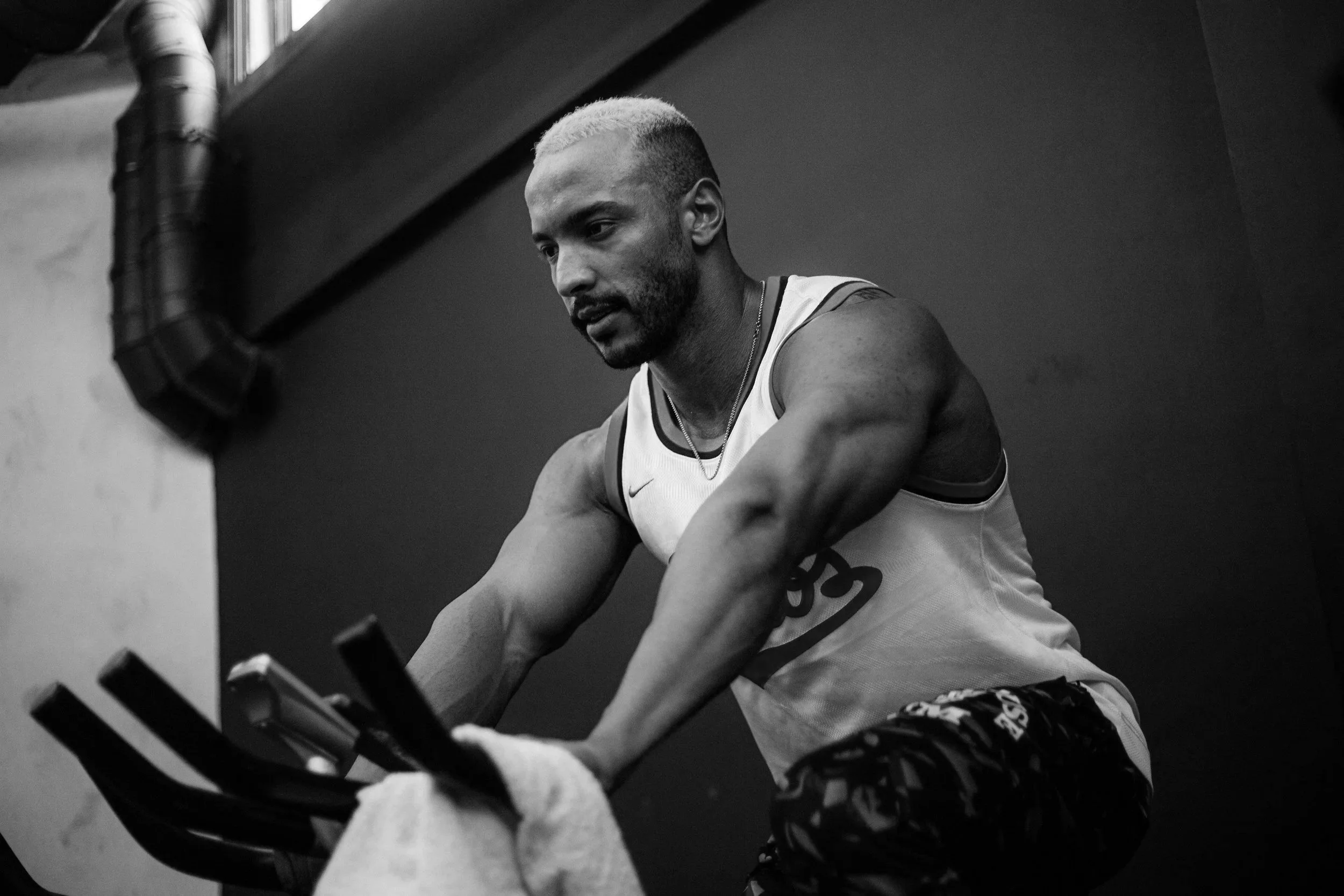 A man with dyed blonde hair and a beard, wearing a sports tank top, is exercising on a rowing machine in a gym.
