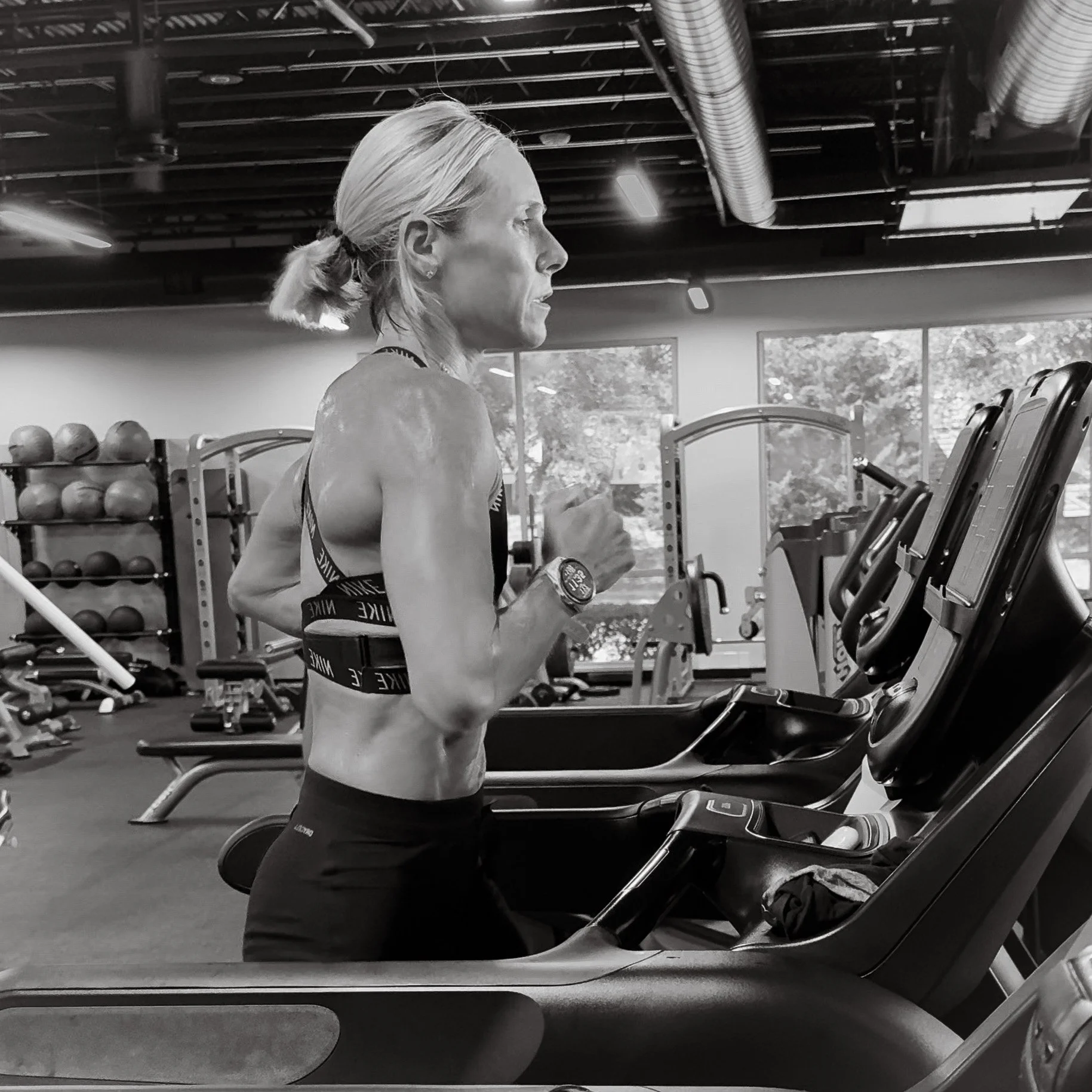A woman with blonde hair in a ponytail running on a treadmill inside a gym, with exercise equipment and large windows in the background.