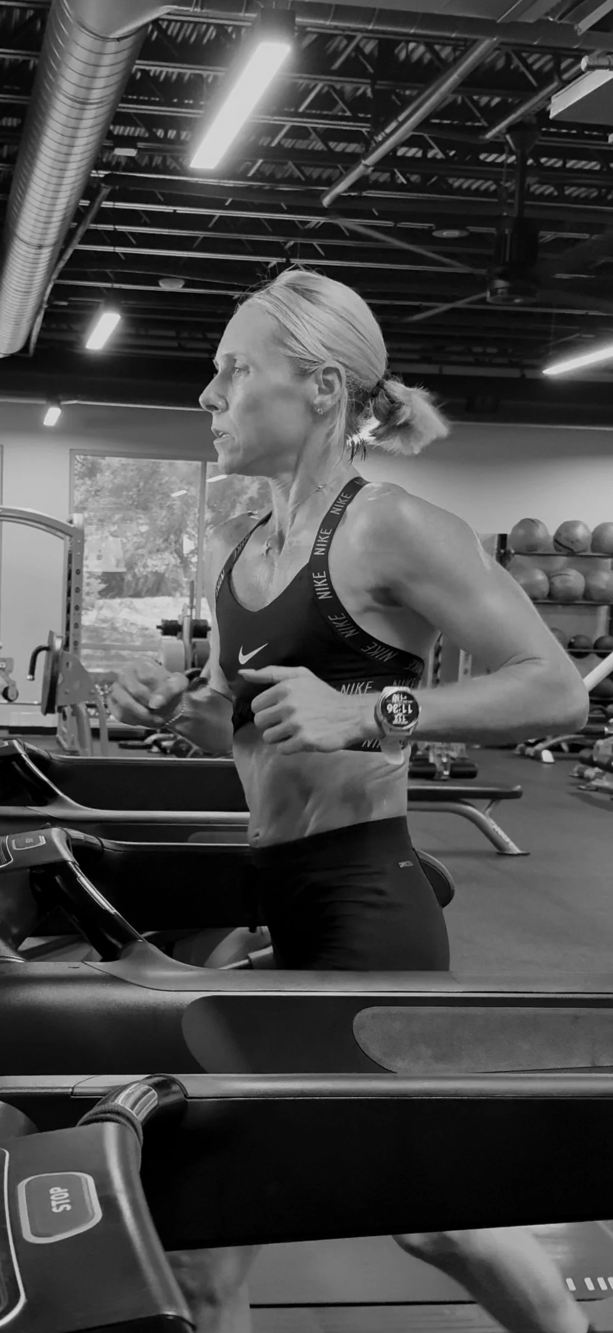 A woman running on a treadmill in a gym, wearing athletic wear and a smartwatch, with gym equipment and kettlebells in the background.