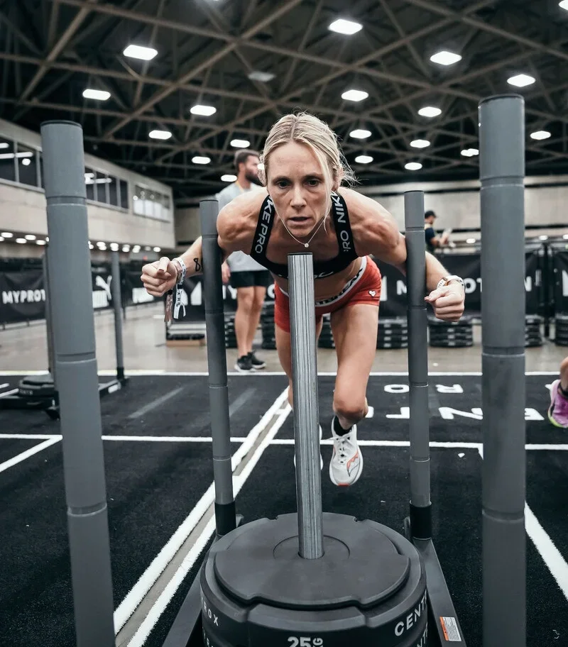 Woman pushing a weighted sled in an indoor gym setting with other people in the background.
