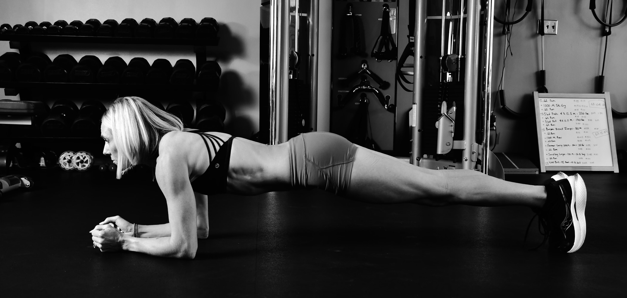 A woman in workout attire performing a plank exercise on the floor of a gym, with workout equipment and a whiteboard in the background.
