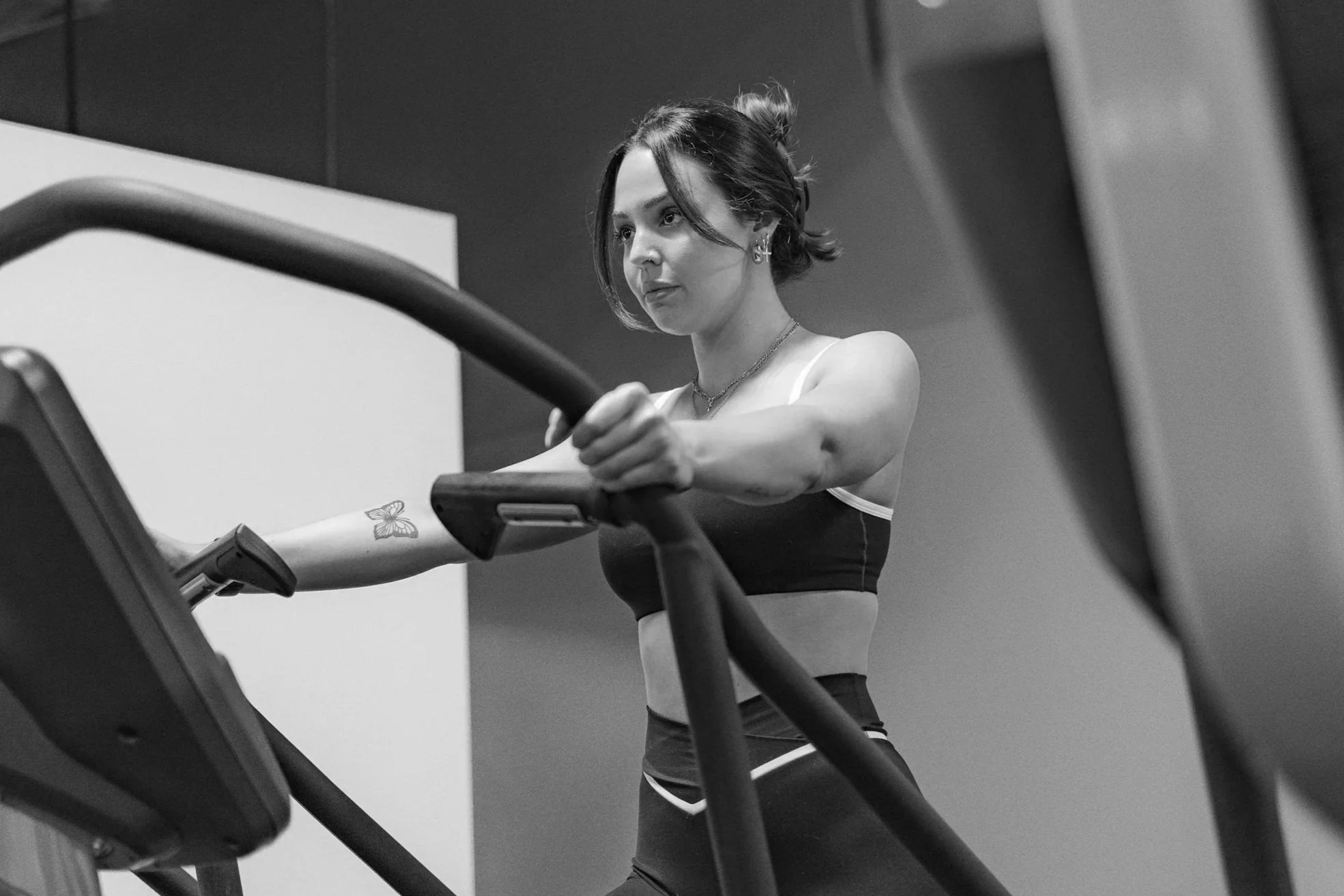 A young woman with a tattoo on her left arm, wearing sportswear, holding onto treadmill handrails, exercising in a gym.