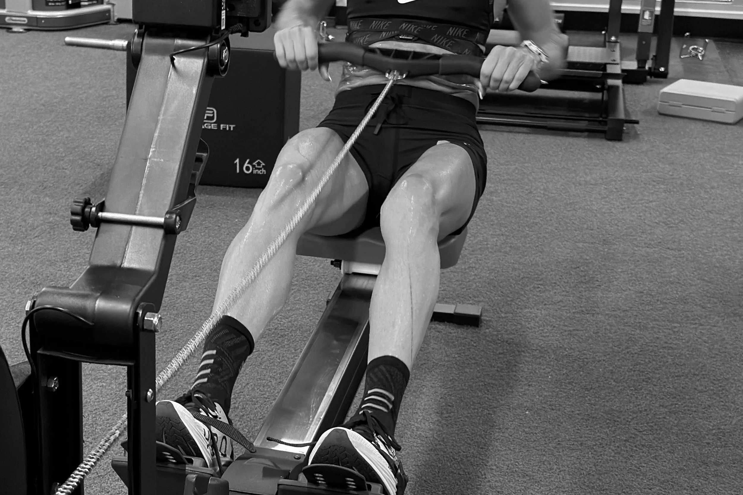 Person sitting on an exercise machine in a gym, holding a handlebar, with legs extended and wearing athletic shoes and shorts.