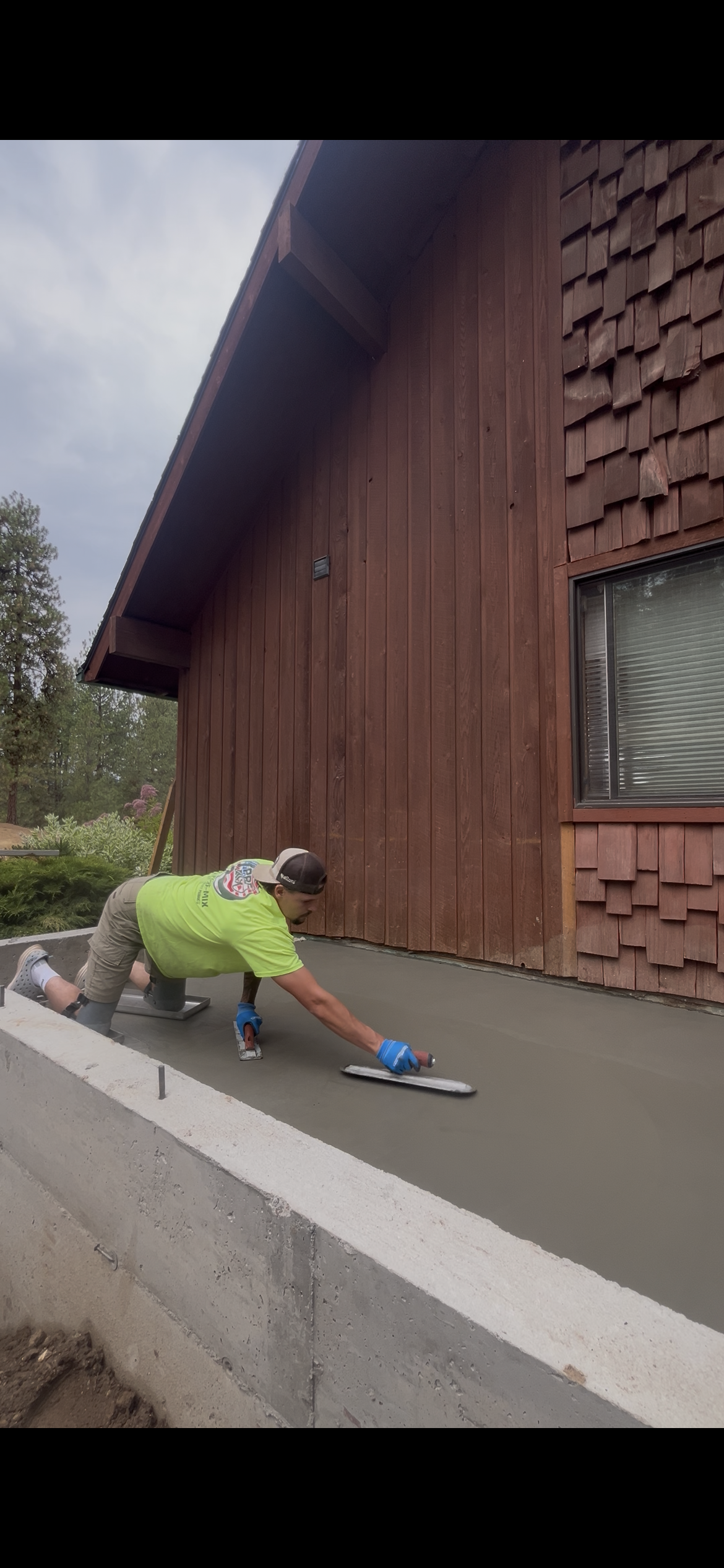 Man kneeling on fresh concrete, smoothing it with a trowel outside a wooden house.