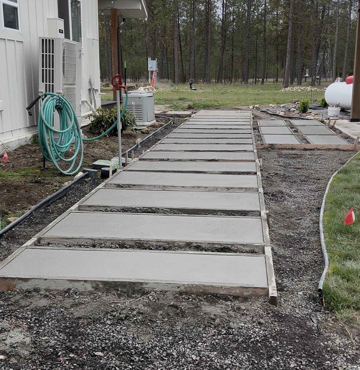 Concrete sidewalk sections being installed in a backyard garden area, with a house on the left and a wooded forest in the background.