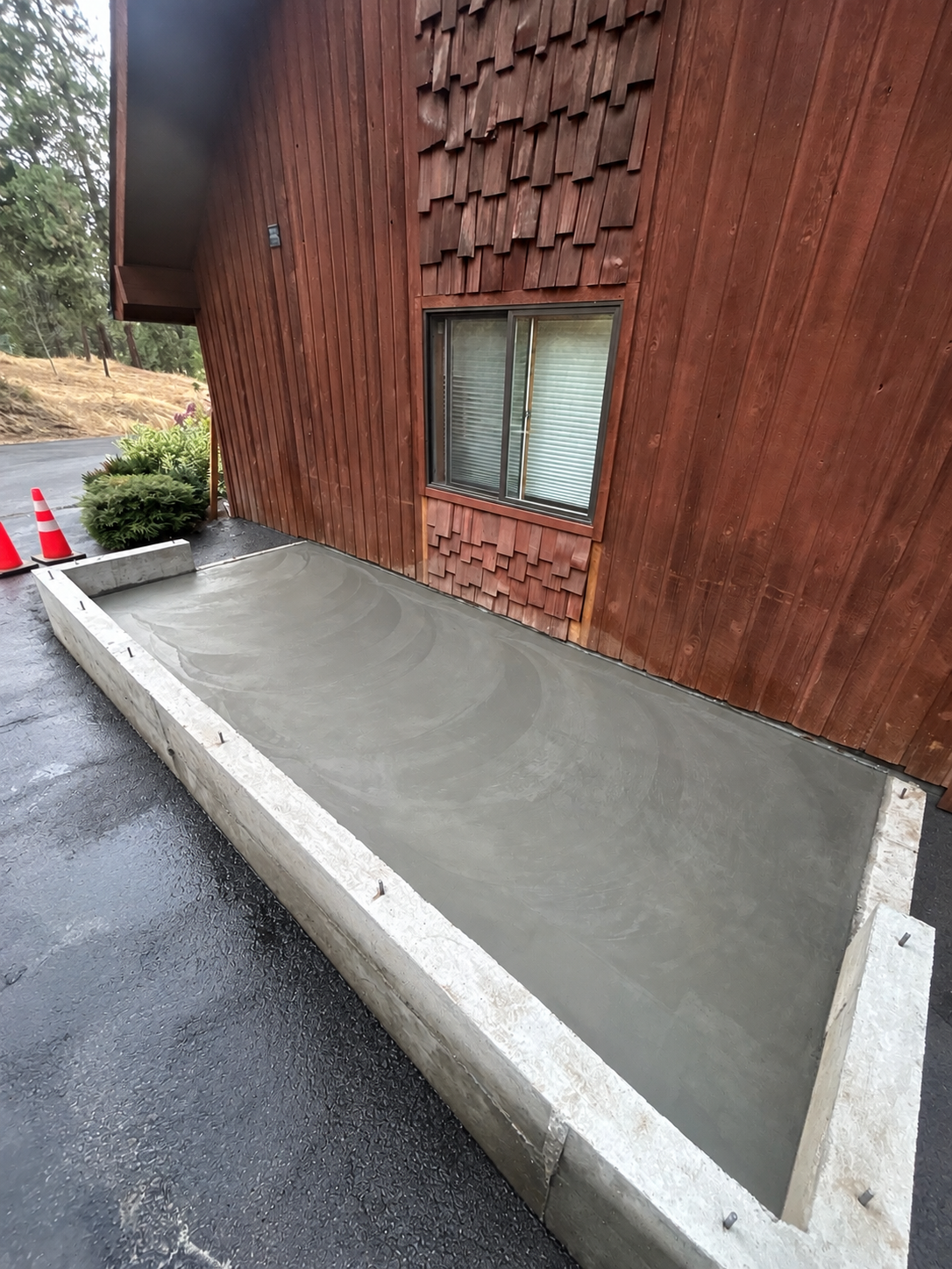 Freshly poured concrete slab in front of a wooden house with a window and red curbside traffic cones.