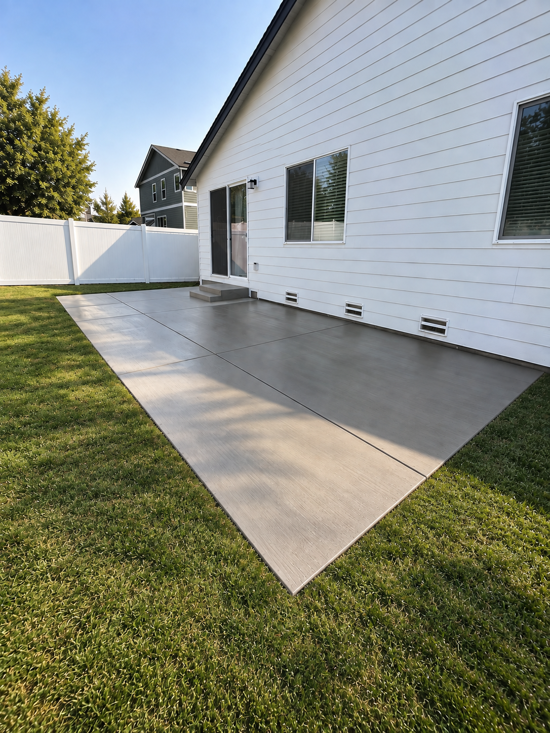 Backyard with a white house, sliding glass door, concrete patio, green grass, tall trees, and neighboring houses in the background.