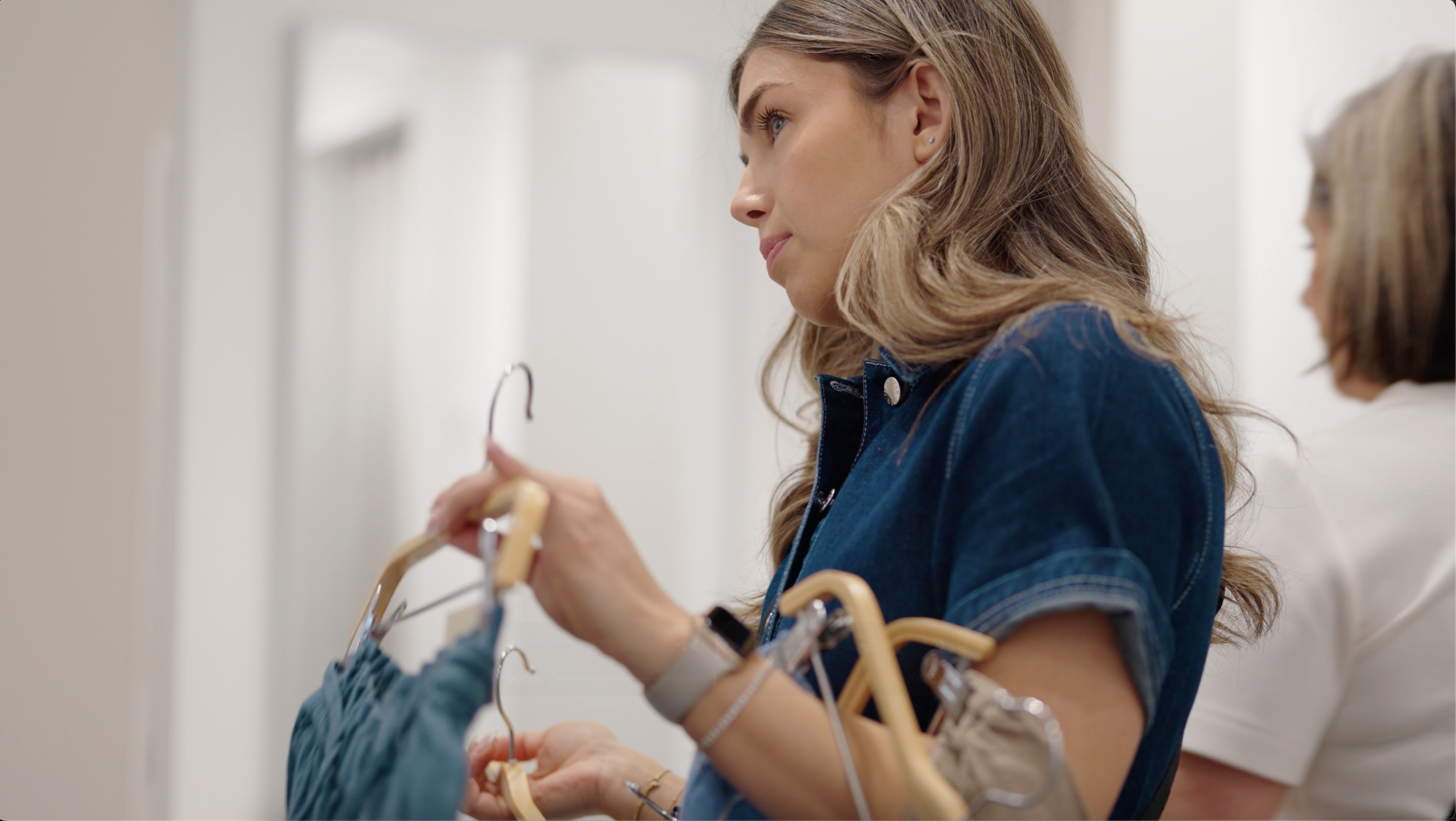Production vidéo événementielle à montréal pour le Basement - Une femme aux cheveux longs et blonds, portant une veste en denim, est dans une cabine d'essayage, essayant des vêtements sur un cintre en bois et en plastique.