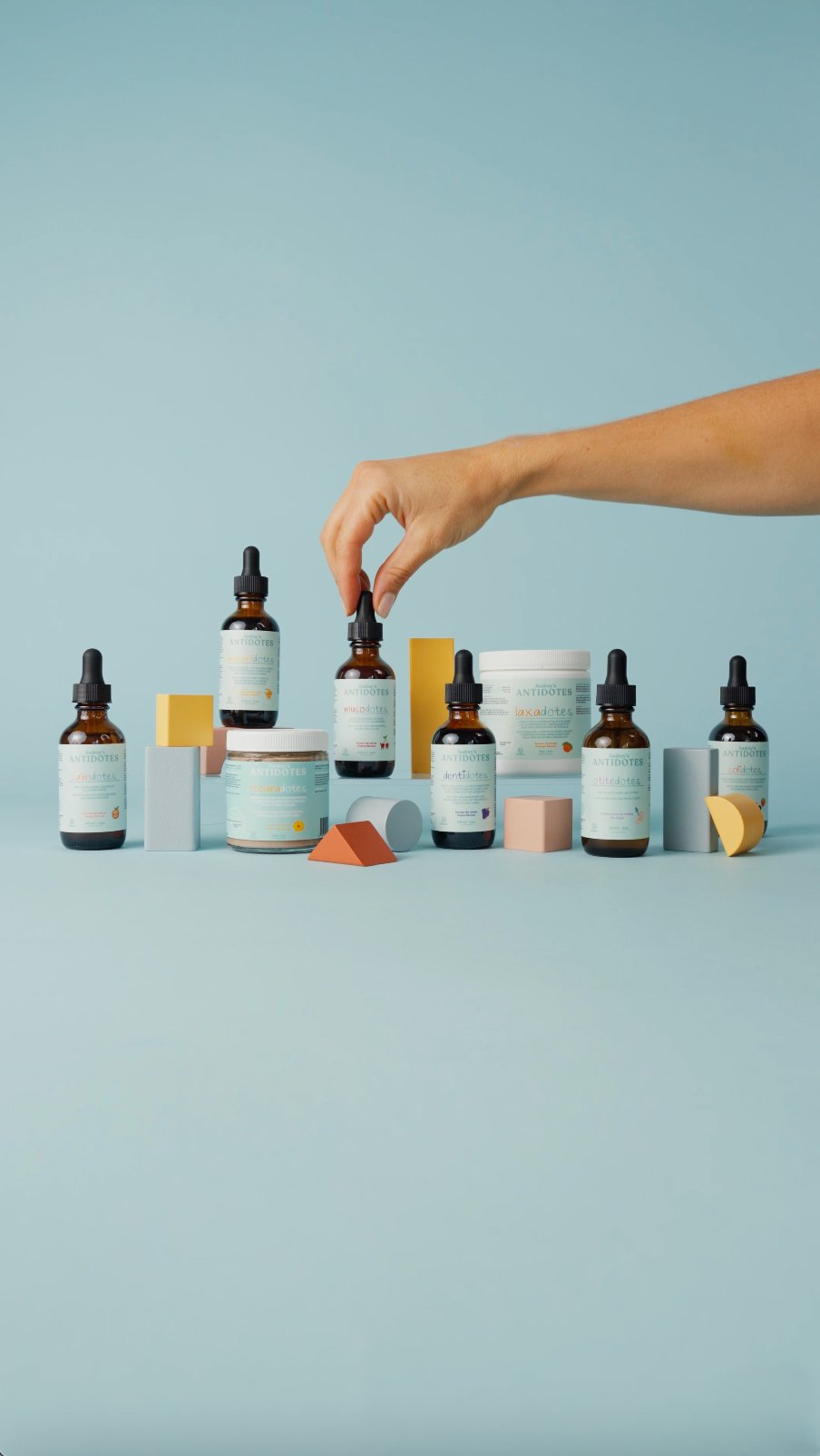 Production et présentation de produits cosmétiques ou de soins, avec plusieurs flacons et contenants disposés sur une surface pastel, une main réglant un flacon entre plusieurs formes géométriques colorées.