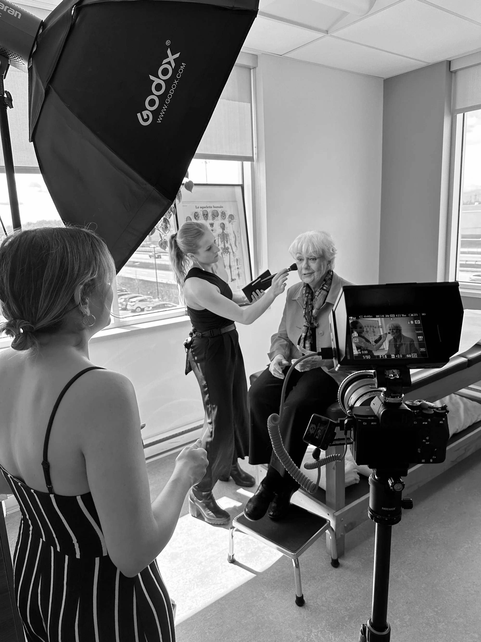 Une séance de tournage à Montréal dans une pièce lumineuse avec une grande fenêtre, où une femme âgée est assise devant un fond blanc, une autre femme jeune la prend en photo, et une maquilleuse en arrière plan.