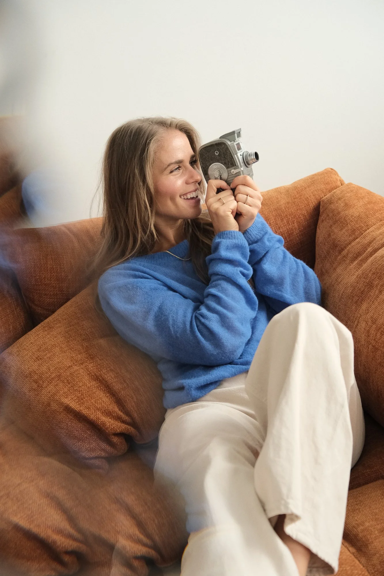 Une femme souriante assise sur un canapé en train de prendre une photo avec un appareil photo vintage.