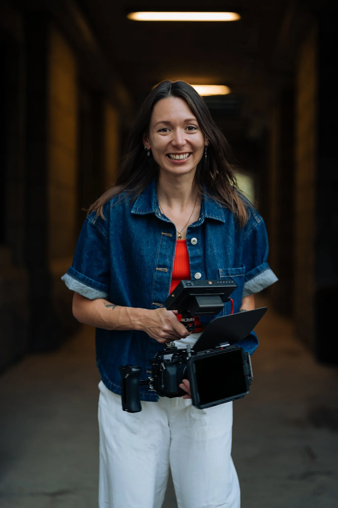 Maggie Parisé tenant une caméra vidéo professionnelle, portant une veste en jean et un pantalon blanc, dans un environnement sombre avec un éclairage au plafond.