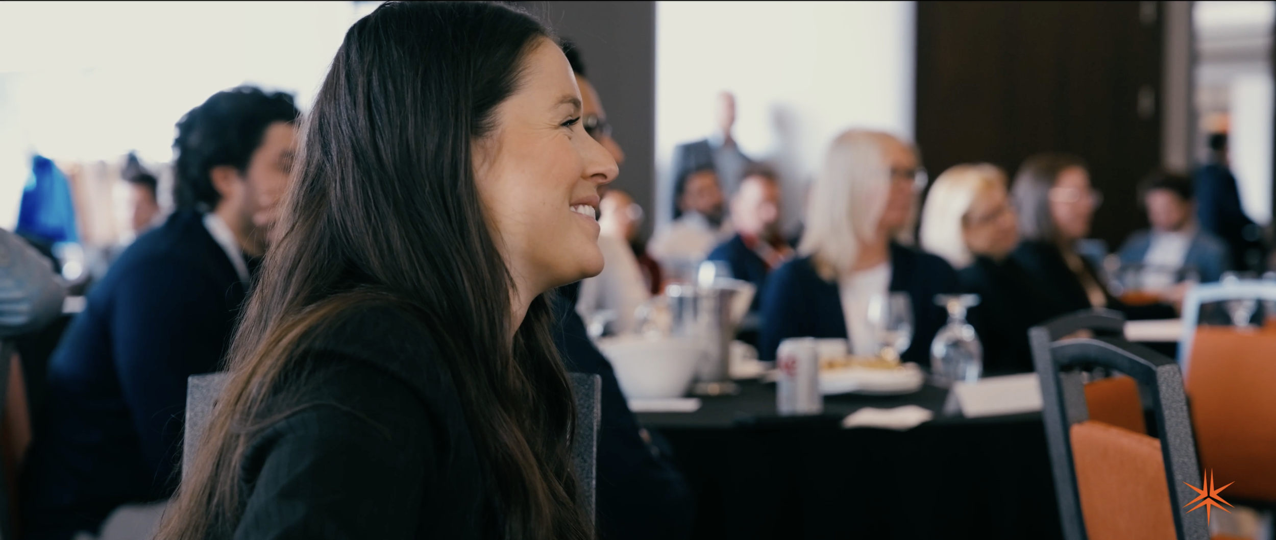 Une femme souriante à une conférence avec d'autres personnes en arrière-plan, assises à une table