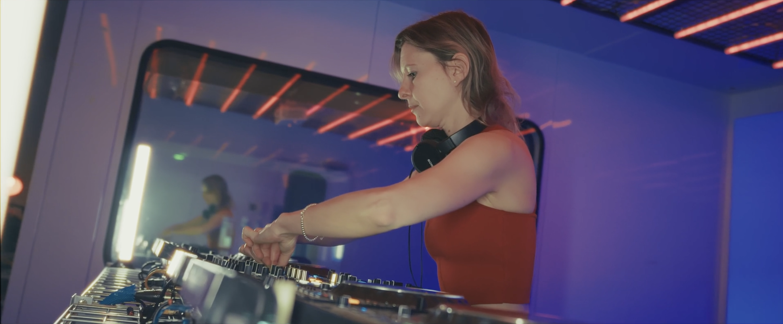 Une femme DJ en tenue rouge, portant un casque, manipule des platines dans une ambiance de club avec éclairages colorés.