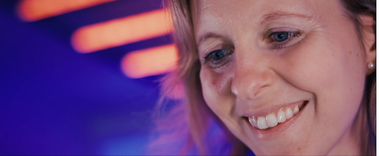 Photo d'une femme souriante avec de longs cheveux bruns, éclairée par des lumières colorées en arrière-plan.