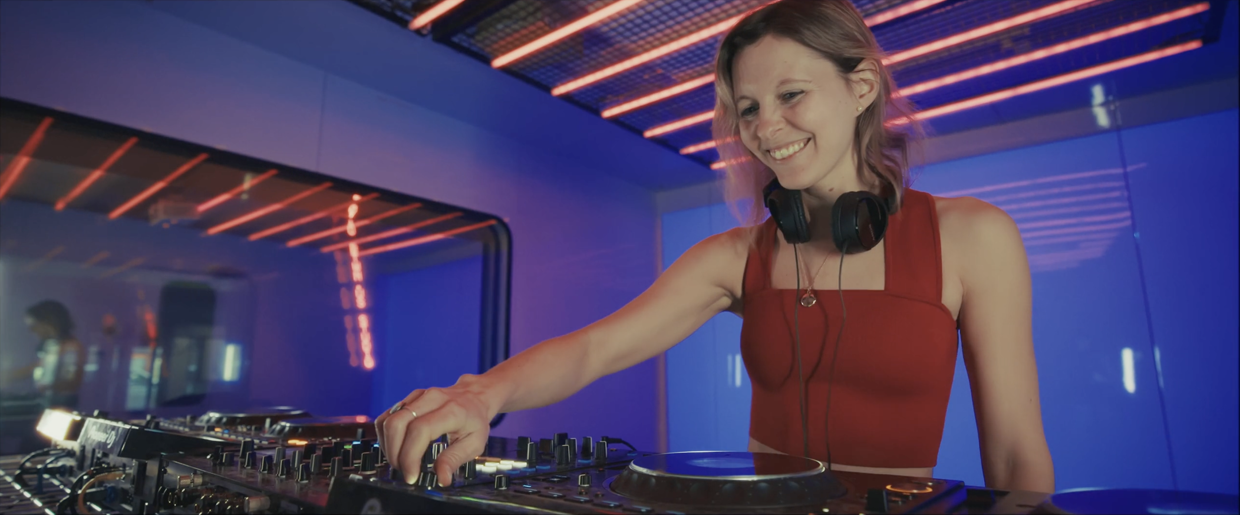 Une femme DJ souriante en tenue rouge, portant un casque, manipule des platines dans un environnement avec des lumières colorées et un décor moderne.