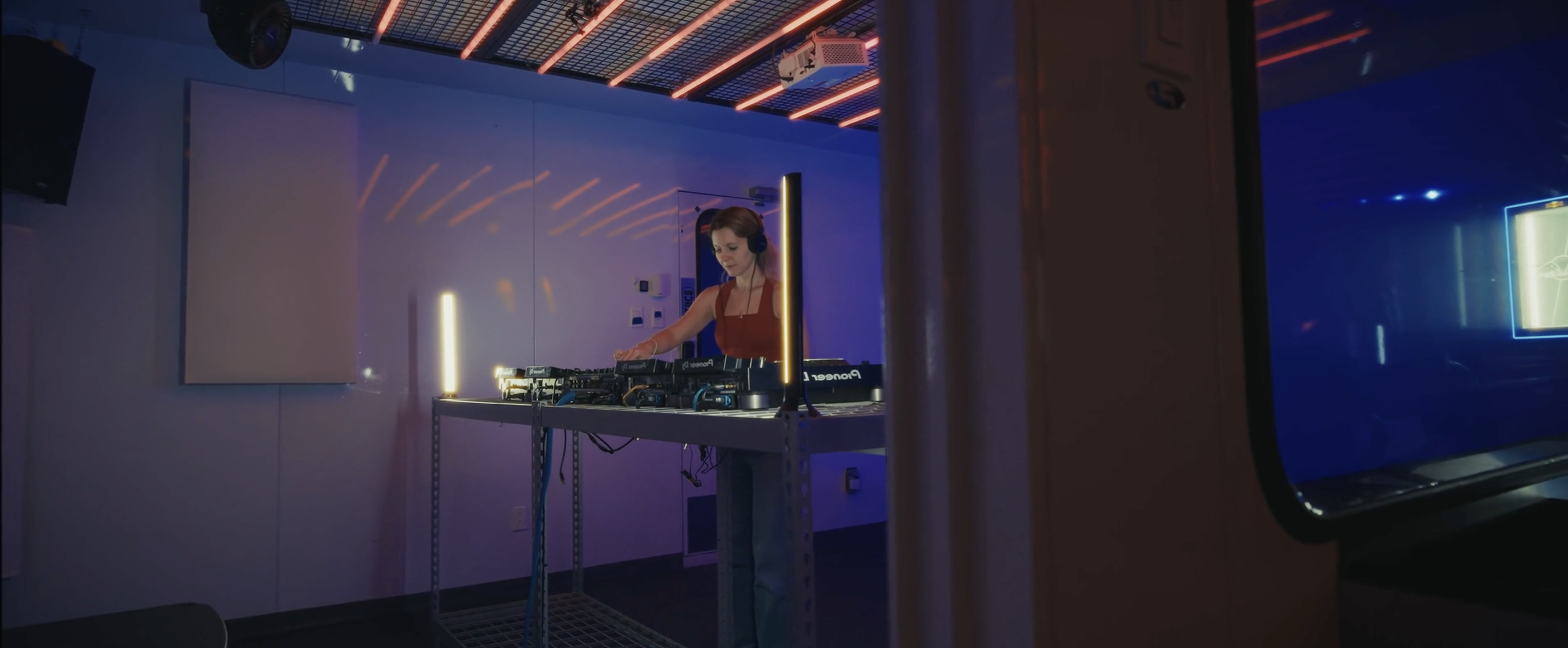 Une femme DJ portant un haut rouge et un casque, procédant à un mixage musical à l'intérieur d'un studio ou d'une salle de fête avec un éclairage néon, vue partielle d'une porte ou d'une cloison en bois de l'autre côté.