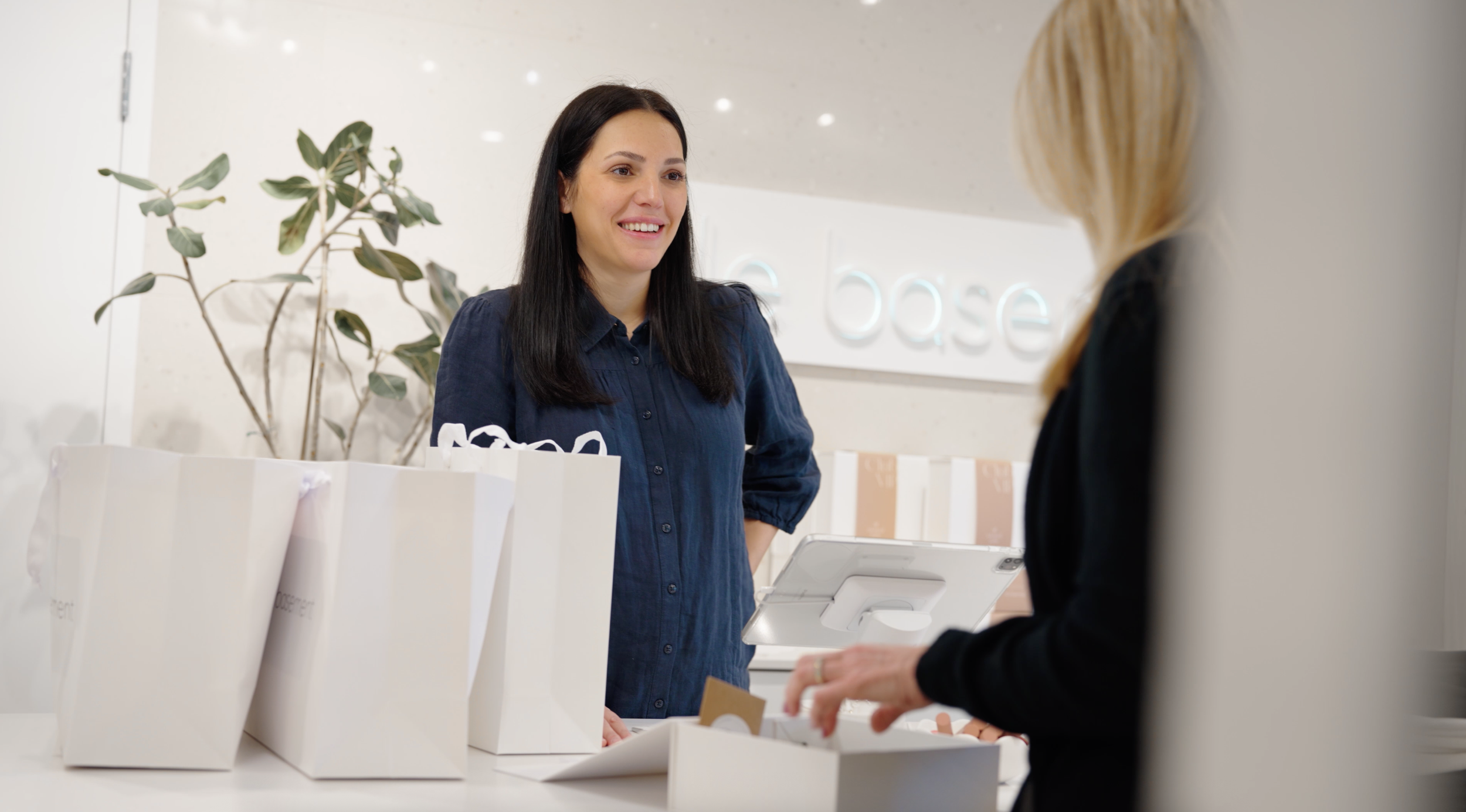 Une femme avec des cheveux noirs, portant une chemise bleue, parle avec une autre personne dont seule une partie est visible. Elles se trouvent derrière un comptoir avec des sacs en papier blanc. Il y a une plante verte derrière la femme et un pannea