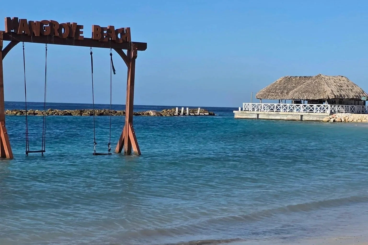 A view of the beach at Manrogye Beach with a wooden swing set hanging over the water in the foreground, a thatched-roof hut on the right, and a small pier in the distance under a clear blue sky.