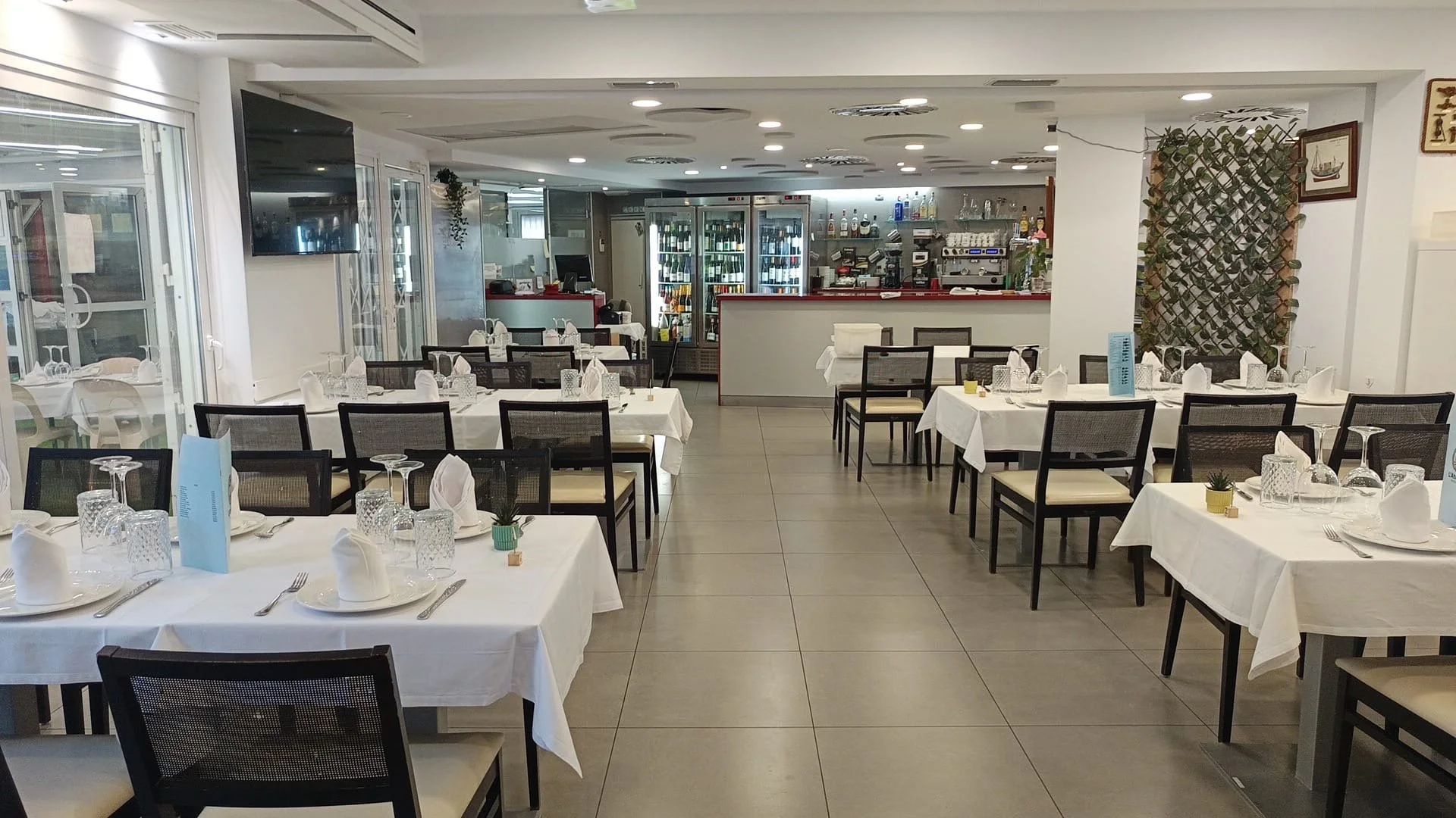 Salón de restaurante con mesas cubiertas con manteles blancos, cubiertos, copas y pequeñas plantas decorativas. En el fondo, barra con refrigeradores y una pequeña área de cocina.