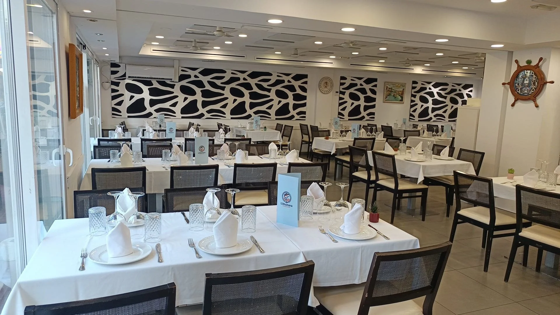 Un restaurante con mesas arregladas con manteles blancos, cubiertos y cristalería, decorado con paredes con diseño de jirafa y algunos cuadros, y ventanales grandes que dejan entrar luz natural.