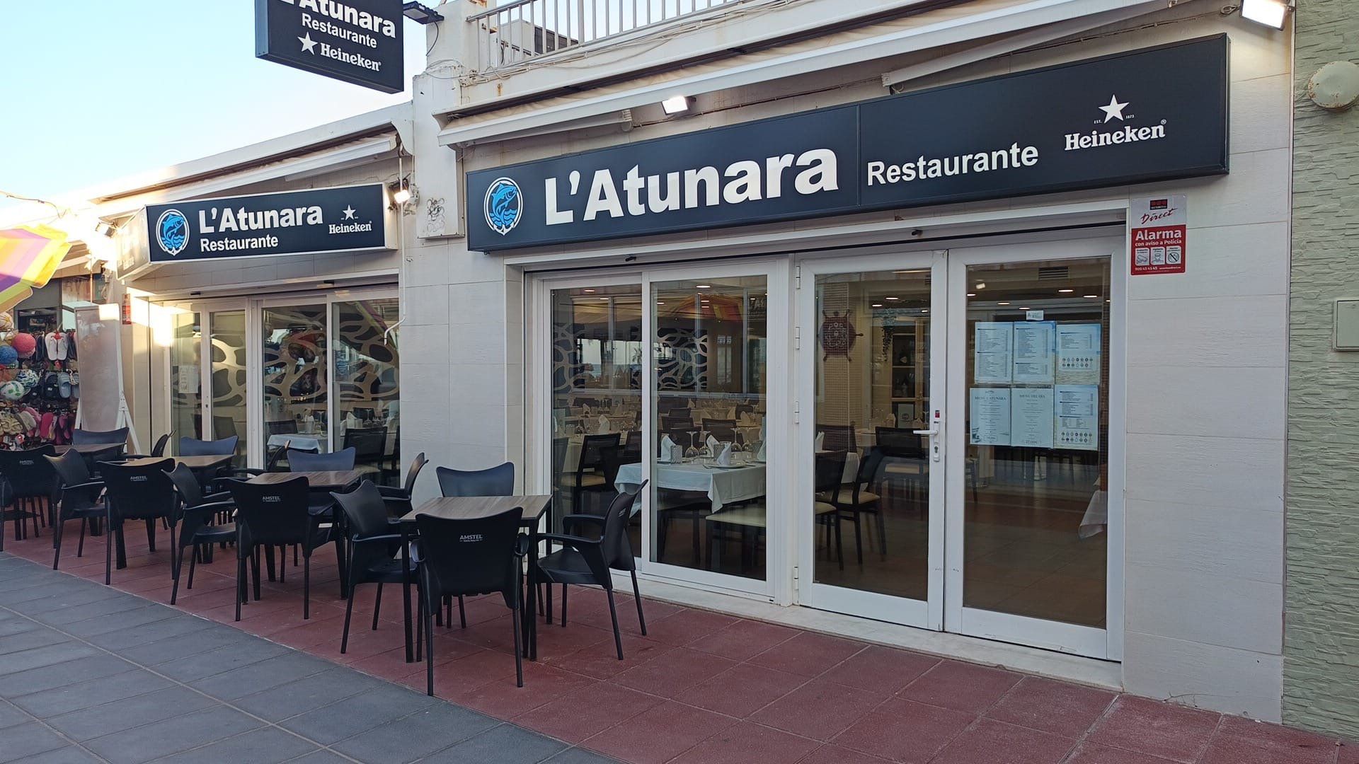 Vista exterior de un restaurante llamado 'L'Atunara', con mesas y sillas en la terraza, y letreros que indican que es un restaurante Heineken. La fachada tiene ventanas grandes y un cartel con el menú visible desde afuera.