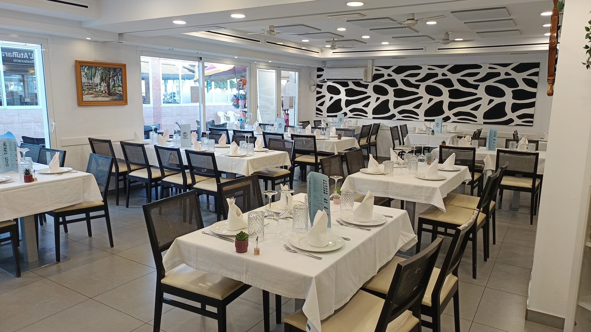 interior de un restaurante con mesas cubiertas con manteles blancos, platos, cubiertos y vasos, y decoración moderna con un muro con patrón negro y blanco