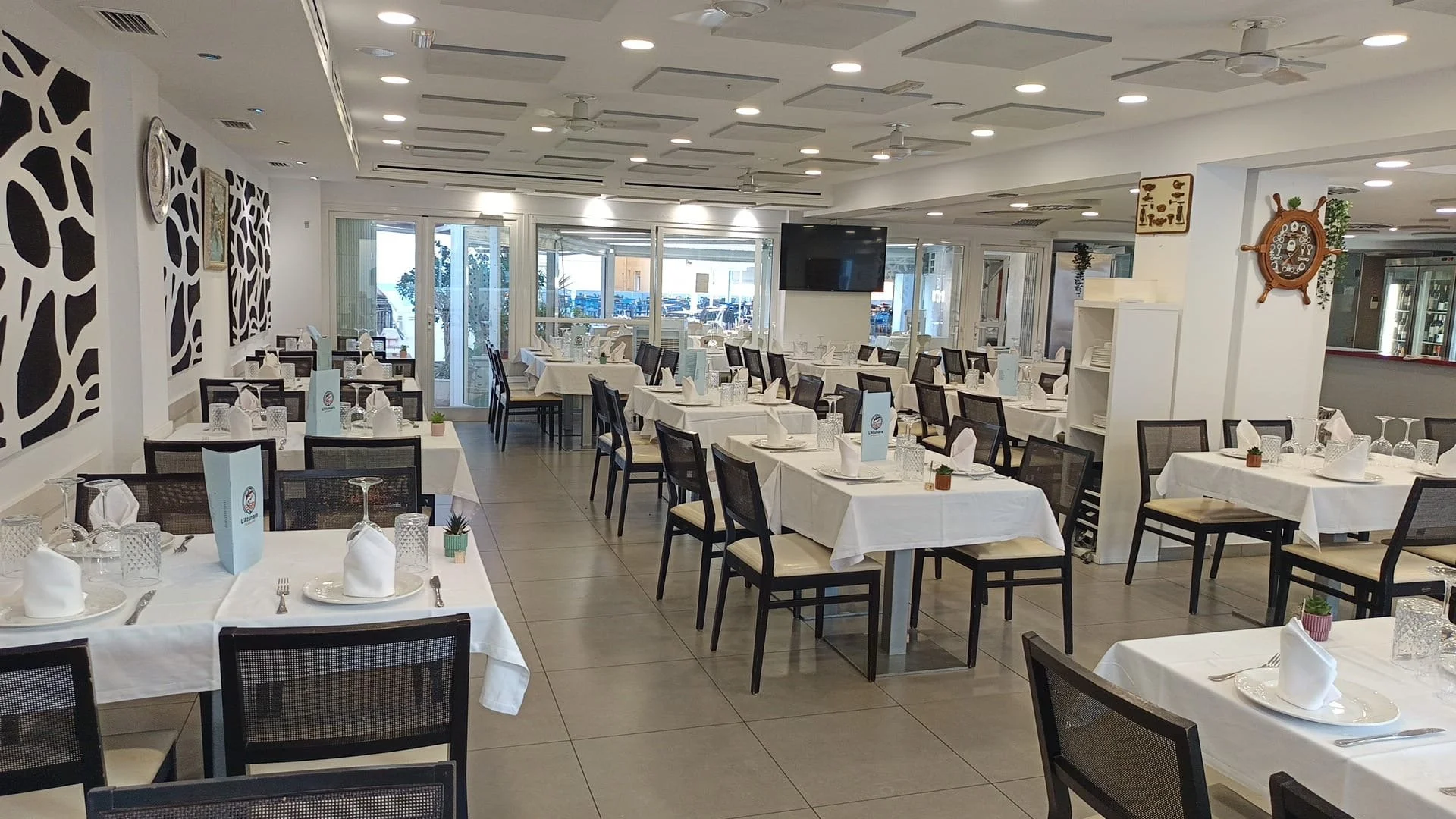 Salón de restaurante elegante con mesas cubiertas con manteles blancos, puesta en mesa con copas y platos, y decoraciones pequeñas. Ventanas que ofrecen vista al exterior con sillas apiladas. Decoración moderna y ambiente luminoso.