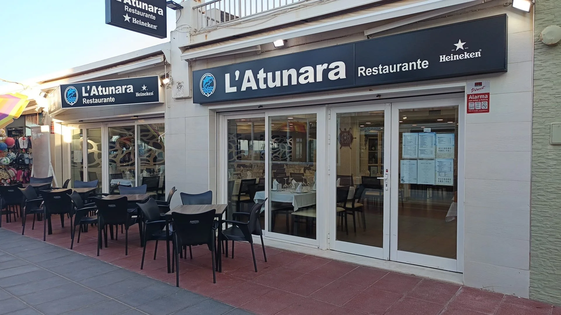 Exterior de un restaurante llamado L'Atunara con áreas de comedor al aire libre, ventanas grandes, y señalización que indica que sirve comida y tiene la marca Heineken.